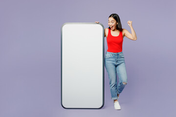Full body amazed young woman of Asian ethnicity wear casual clothes red tank shirt big huge blank screen mobile cell phone with area do winner gesture isolated on plain pastel light purple background.