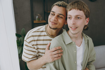 Young dreamful calm happy minded couple two gay men wearing casual clothes together looking at window at home flat rest spend free spare time in living room. Pride day june month love lgbtq concept.