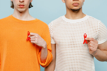 Cropped young couple two gay men wearing casual clothes together hold AIDS awareness red ribbon isolated on plain light blue color background studio portrait. Pride day june month love LGBTQ concept.