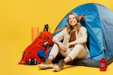 Full body smiling fun young woman sit near bag with stuff tent look camera isolated on plain yellow...