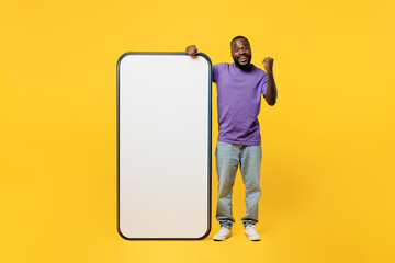 Full body young man of African American ethnicity wear casual clothes purple t-shirt big huge blank screen mobile cell phone smartphone with area do winner gesture isolated on plain yellow background.