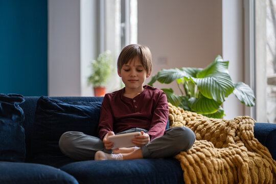 Little boy kid playing mobile games while resting at home, child sitting on sofa with smartphone in hands. Schoolboy staring at phone screen. Children and gaming addiction concept