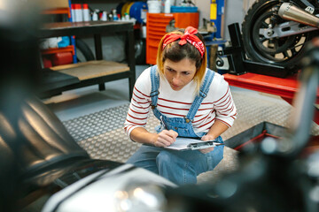 Concentrated mechanic woman with clipboard reviewing damages in motorbike on garage