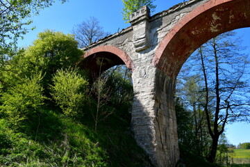 A close up on an old, abandoned and damaged tran bridge made out of concrete, red brick and other materials located in the middle of a field surrounded with hills, forests, and moors seen in Poland