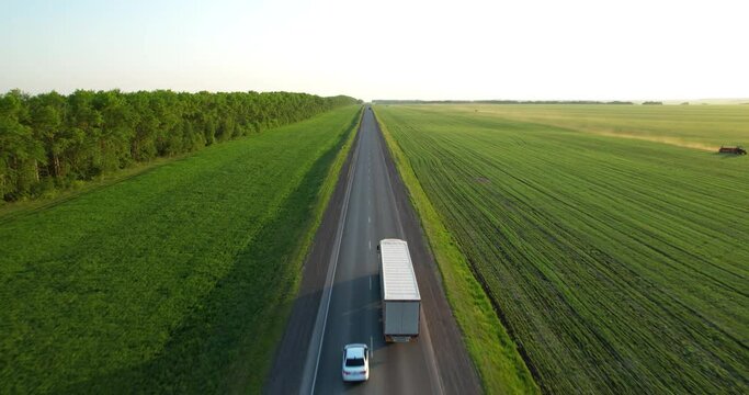 Trucks with a semi-trailer drive along the highway, aerial shot. Freeway truck traffic at sunset in summer. Green field. Drone view