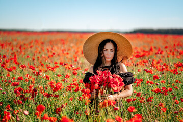Portrait of a beautiful brunette woman in a poppy field. She's wearing a big hat and a basket of red flowers