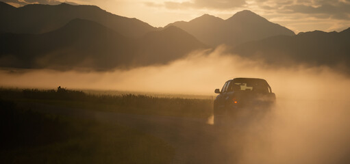 Pickup truck car drives fast on the country road with clouds of dust and beautiful mountains in the background. Sunrise in the valley with steam. Panoramic view with free space for text - obrazy, fototapety, plakaty
