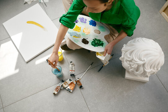 Top view on young woman artist mixing colors for painting while sitting on floor