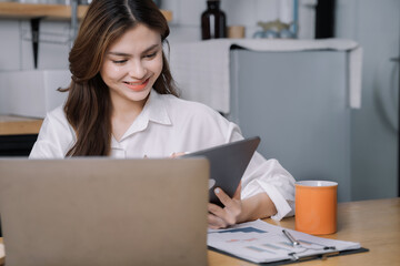Fototapeta premium Young beautiful mixed-race girl using laptop and smartphone communicating internet with home clients, reusable coffee mugs on comfortable desk, working remotely, e-learning concept.