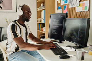 African American programmer in wireless headphones developing computer codes on computer sitting at...