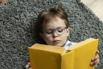little boy with glasses reading a book lying on the floor