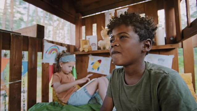 Slowmo of African American elementary age boy blowing bubble with gum while Caucasian boy of same age playing with tablet at background of treehouse