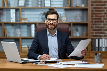 Fototapeta na wymiar Portrait of a young handsome man, freelancer, businessman, accountant. Sitting in the office, looking at the camera, smiling. Works on a laptop, with documents, fills in forms with a pen