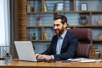 Successful mature businessman at work with laptop inside office, man in business suit sitting at desk typing on computer, satisfied with achievement result and work.