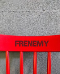 Red chair back on copy space concrete wall with text - FRENEMY - person who combines the characteristics of friend and enemy, rival pretends to be a friend