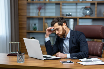 Obraz premium sad and depressed businessman in depression working inside modern office building, man in business suit with glasses and beard using laptop for work, boss disappointed with work result