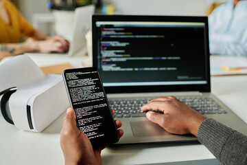 Close-up of man writing security codes on gadgets and connecting smartphone to laptop sitting at his workplace