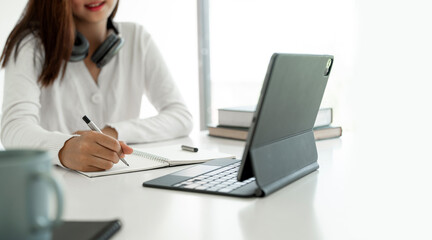 Young woman using digital tablet for online learning at home, writing on her notebook.