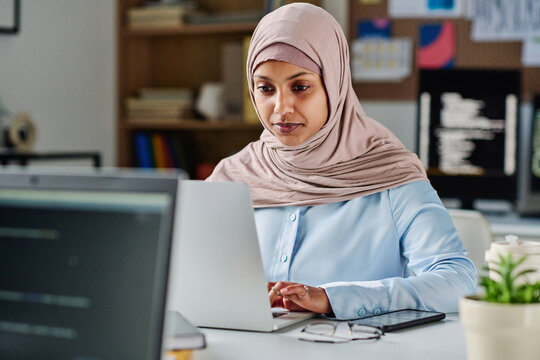 Young muslim woman working online on laptop while sitting at her workplace in IT office