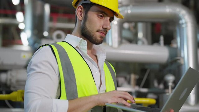 Professional male electrical engineer in safety uniform working on laptop at factory site control room. Industrial technician worker maintenance and checking power system at manufacturing plant room.