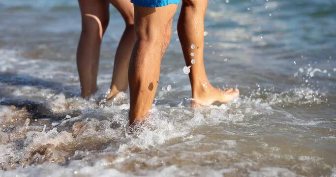 People kick rolling sea waves with bare feet at resort on beach. Active tourists enjoy day on beach. Couple vacation and entertainment on seaside slow motion