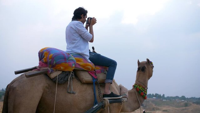 Indian boy sitting on camel's back and capturing photos - photography  hobby  Pushkar mela  sand dunes  sand safari  camel safari. An attractive male in casual clothes sitting on a camel at the Mel...