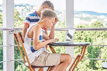Brother siblings look together at difficult game on grey laptop on hotel balcony. Preteen schoolboy tries to win serious game during summer holidays