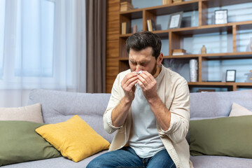 Sick mature man sitting on sofa at home, man has allergy and runny nose, sneezing in living room alone.