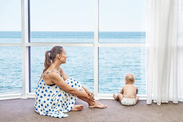 Happy mother plays with toddler son against azure sea view from hotel room window. Boy enjoys looking at new things on sea holidays in summer