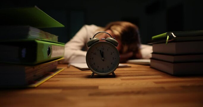Alarm clock stands on wooden desk against sleeping lady office worker. Tired female rests at workplace feeling exhaustion from working at night