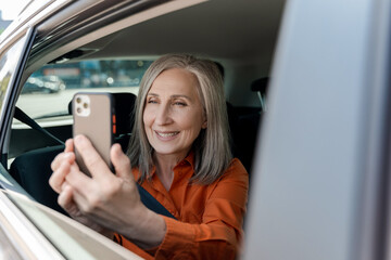 Smiling attractive senior woman holding mobile phone, taking picture looking at window window sitting inside taxi. Travel, vacation, Transportation concept