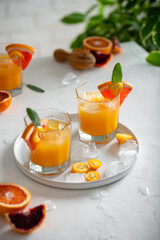 Glasses of freshly squeezed juice with leaves and fruits on table on bright background. Grapefruits, oranges, bloody oranges, kumquats and ice cubes.