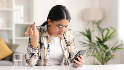 Worried young Indian girl sitting at the table looking at her phone and does not understand why it is not working. The student adjusts her glasses because she does not believe the bad news she saw.