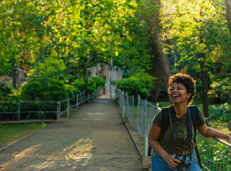 Happy young black African American woman photojournalist with dark curly hair laughing happily with open mouth looking at friend with photo camera hanging around neck in green park at sunset