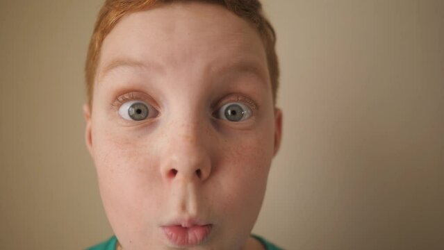 Little red-haired boy looking into camera with wow expression indoor. Portrait of ginger child with freckles showing surprise and amazement. Close up shocked male face of small kid inside. Slow motion