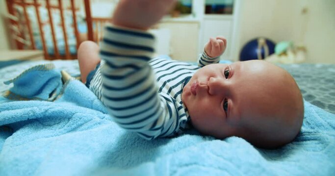 An active three-month-old boy lies on his back, looks into the camera. Healthy baby in a striped bodysuit on the bed in the bedroom. Handsome white American boy.
