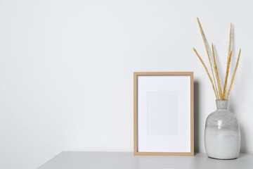 Empty photo frame and vase with dry decorative spikes on white table. Mockup for design