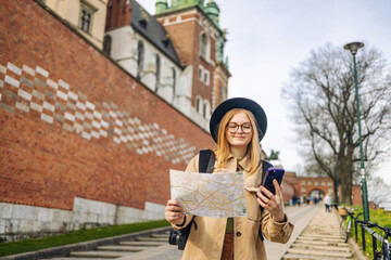 Stylish clothes blond girl tourist travels looking for directions on map and phone while traveling through the old town of Krakow in spring, Poland. Spring view of Wawel Royal Castle 