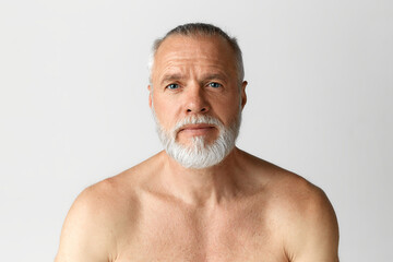 Portrait of handsome middle-aged man with blue eyes posing shirtless, looking at camera against grey background. Well-kept skin. Concept of male beauty, face and skin care, daily procedures, age