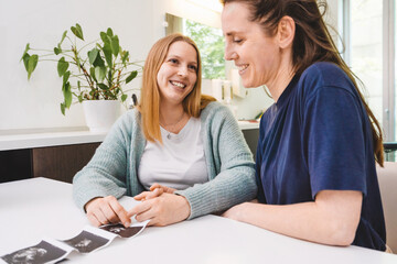 A joyful caucasian lesbian couple, holding hands while sitting at the gynecologist's desk, with their baby's ultrasound visible on the table. - obrazy, fototapety, plakaty