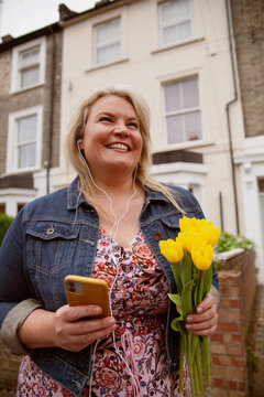 Happy plus-sized woman holding flowers while video-chatting on her phone in a neighborhood