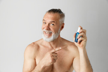 Portrait of smiling, handsome, mature man with gray hair and beard posing with face toner against grey background. Concept of male beauty, face and skin care, daily procedures, age. Ad