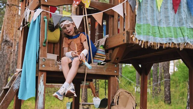 Full shot of Caucasian elementary age boy sitting in treehouse pretending to shoot with badminton racket