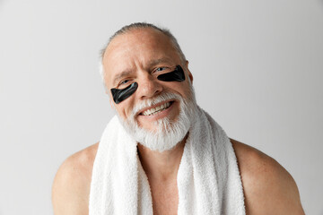 Fototapeta premium Portrait of handsome mature man with towel, shirtless smiling, posing with under-eye patches against grey background. Concept of male beauty, face and skin care, daily procedures, age