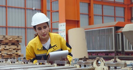 Young latin technician women in safety hard hat holding clipboard working and checking stock while walking at industrial factory