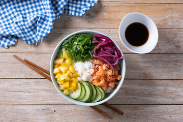 Poke bowl with rice, salmon,cucumber,mango,onion,wakame salad, poppy seeds ands sunflowers seeds on wooden table