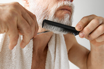 Barber. Cropped image of mature man's face, male model taking care after face, cutting and brushing beard against grey background. Concept of male beauty, face and skin care, daily procedures, age