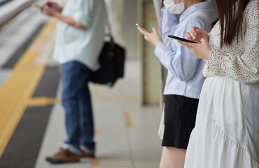 駅のホームで携帯電話を見ながら電車待ちの人々の姿