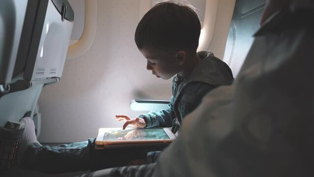 Cute boy sitting in an airplane plays a tablet computer. Entertainment modern technology. Travel concept. Mobile games for children. The child watches cartoons with a tablet.