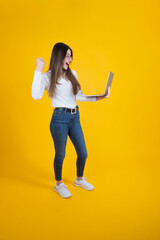 Celebrating success, full body length cheerful woman celebrating success. Caucasian lady raising clenched fist, looking and holding modern laptop. Yellow studio background. Cheering victory.